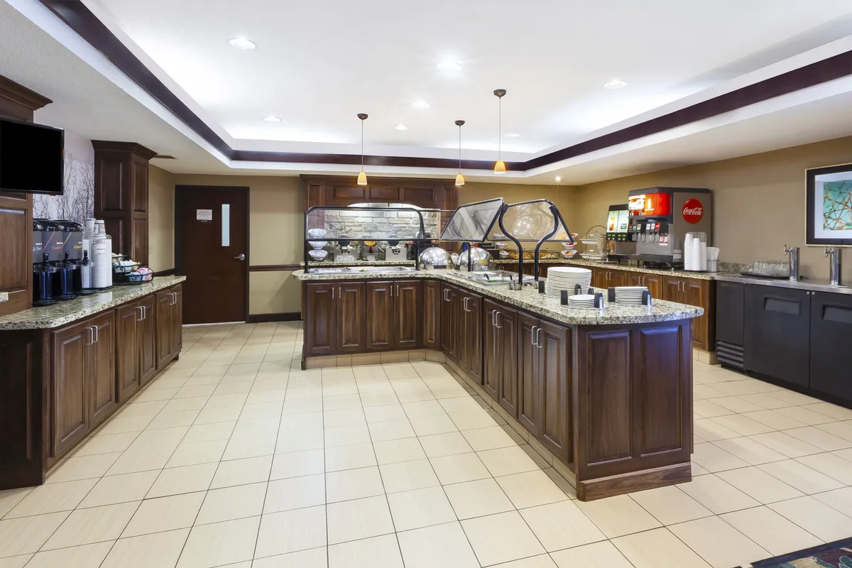This image showcases a well-lit buffet counter featuring a coffee station, a beverage dispenser, and decorative artwork. The space is inviting, with a modern communal breakfast area vibe, highlighting the option for guests to enjoy a complimentary buffet breakfast. The coffee station is clearly visible, supporting the narrative of waking up to fresh coffee.