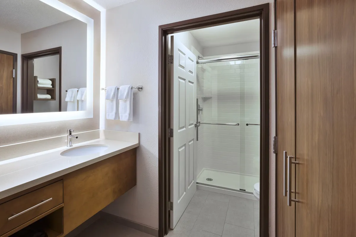 A modern bathroom featuring a well-lit vanity with a sleek sink, a spacious shower area, and a large mirrored wall. Plush towels are neatly arranged, and complimentary toiletries are visible on the countertop, creating an inviting, spa-like atmosphere.