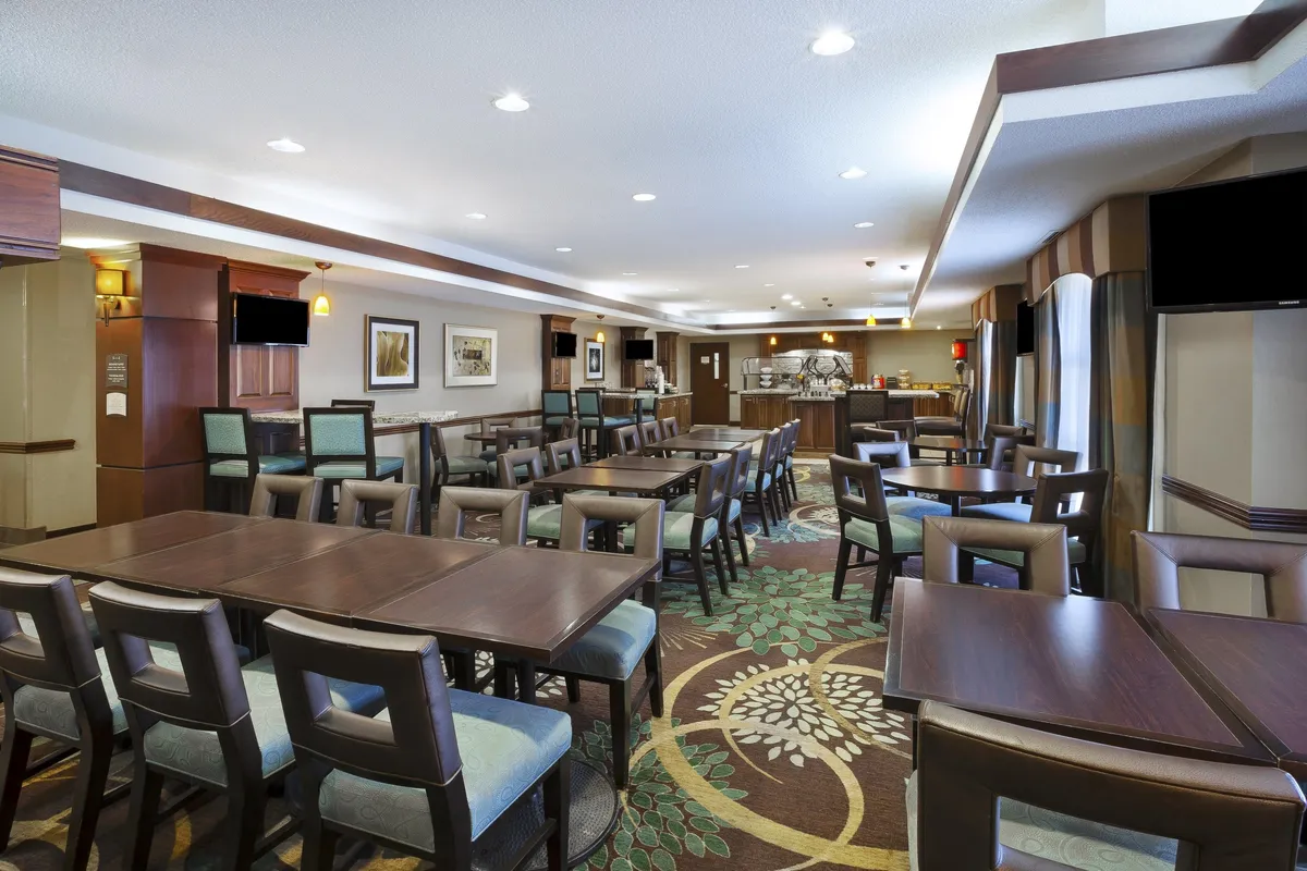 A spacious, warmly decorated dining area featuring wooden tables, comfortable chairs, a decorative carpet, wall art, and a visible service counter. The setting is ideal for intimate meals or group gatherings, aligning with the concept of leisurely in-suite dining and social interaction.