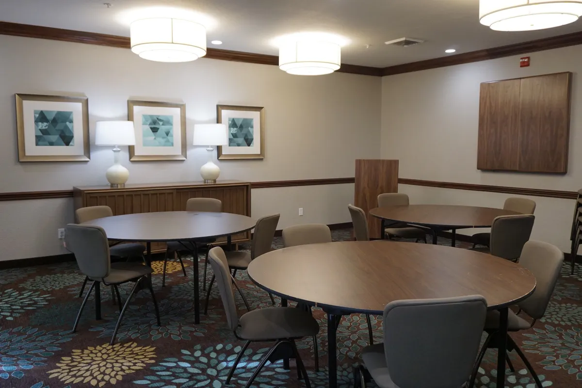 A bright, inviting interior space with circular tables set beneath decorative lamps, surrounded by framed artwork on the walls. The arrangement encourages both relaxation and social interaction, reinforcing a communal, welcoming environment.