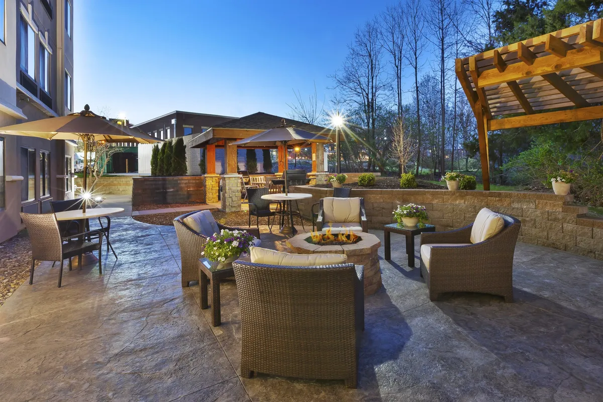 This exterior image showcases a charming outdoor space with a fire pit, ample seating, a pergola, and lush landscaping. Umbrellas and cozy furniture invite social gatherings or relaxing evenings under the stars, providing guests a place to unwind after a productive day. The atmosphere strikes a balance between work and relaxation, embodying the blend of productivity and comfort highlighted in the section.