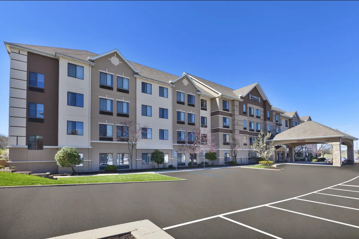 This image captures the main entrance of Staybridge Suites, featuring a covered canopy, large welcoming windows, and a thoughtfully landscaped front area. The scene evokes a sense of arrival and first impressions, suggesting convenience and accessibility for business travelers arriving for meetings or conferences. The outdoor seating area visible in the photo hints at opportunities to relax or connect after a busy workday.