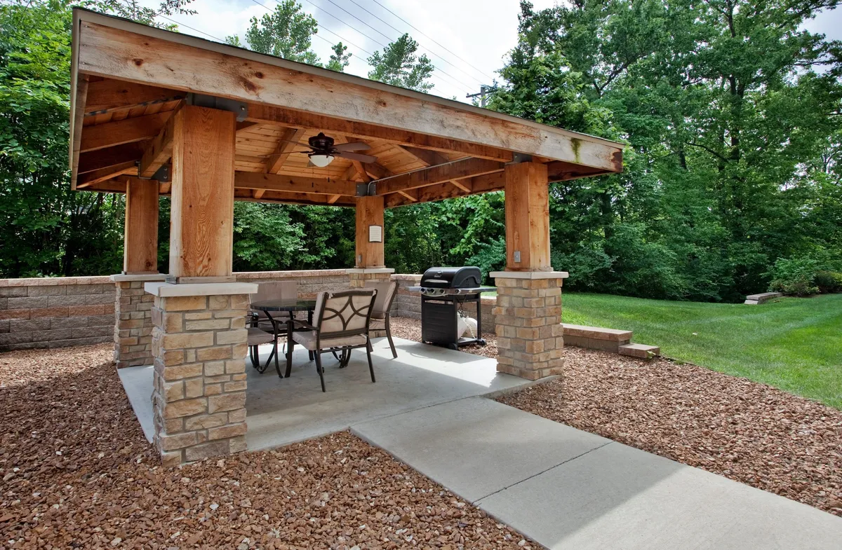 This image shows a welcoming outdoor area featuring a wooden gazebo, a grill, and an outdoor seating arrangement. The space is designed for barbecuing, social gatherings, and relaxing outdoors, with a casual and relaxed ambiance.