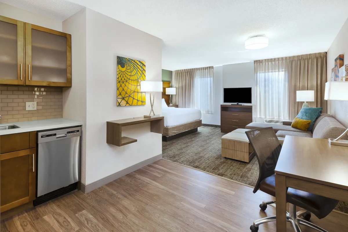 The image showcases a bright, modern studio featuring a cozy bed, a comfortable sofa, a sleek kitchenette, and a wall-mounted TV, all arranged to maximize space and comfort. The inviting setup hints at possibilities for relaxing movie nights, home-cooked meals, or productive work sessions, all within a stylish, well-lit environment.