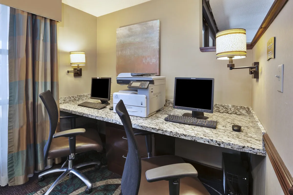 A business center with a granite countertop housing computers, a printer, and elegant lighting. The setting is orderly and professional, ideal for focused work or virtual meetings.