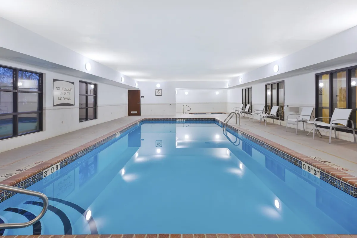 A serene indoor swimming pool with sparkling blue water, surrounded by lounge chairs and large windows allowing in natural light. The space appears clean, modern, and inviting, perfect for swimming, relaxing, or socializing with fellow guests.